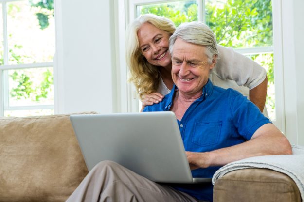 Happy senior couple using laptop while sitting on sofa against window at home Happy senior couple using laptop while sitting on sofa against window at home